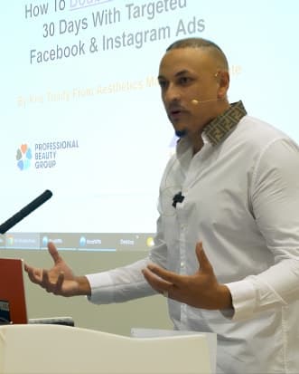 Kris Trinity presenting at a Professional Beauty Group event, gesturing on stage with a headset mic
