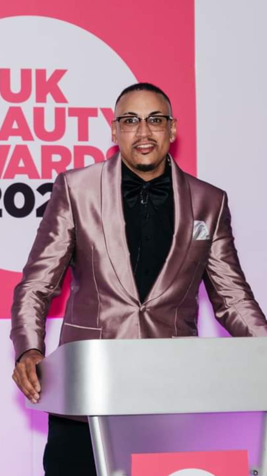 Kris Trinity at a podium at the UK Beauty Awards, wearing a metallic blazer and bow tie